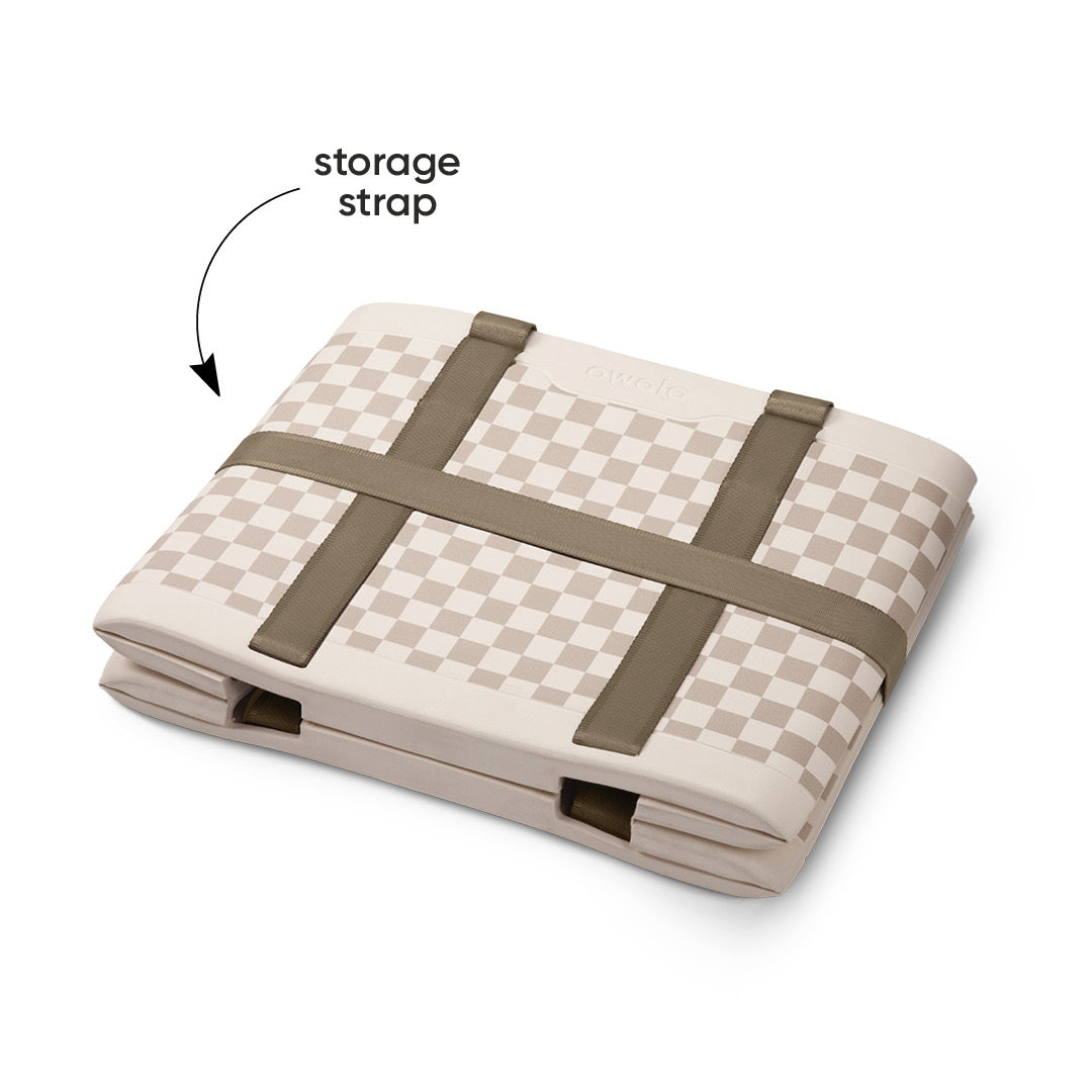 Owala Sun-Day Collapsible Tote in Checkin' In shown folded flat with its storage strap secured across the top, cream and tan checker exterior with olive green storage strap, with text overlay reading "storage strap"